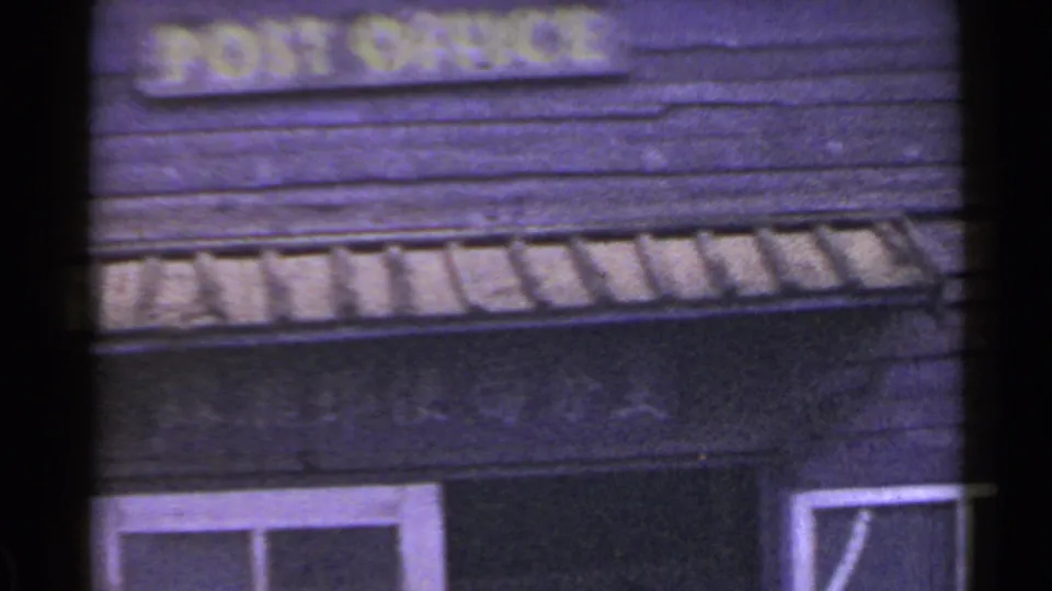 1953 Japan 8mm frame centered on a yellow POST OFFICE sign across dark wooden siding, with roof trim and a lower window visible.