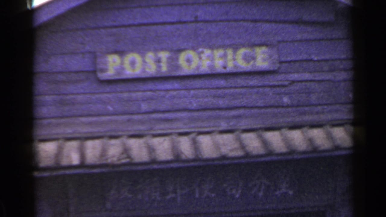 1953 Japan 8mm frame showing a yellow English POST OFFICE sign mounted on dark weathered boards.