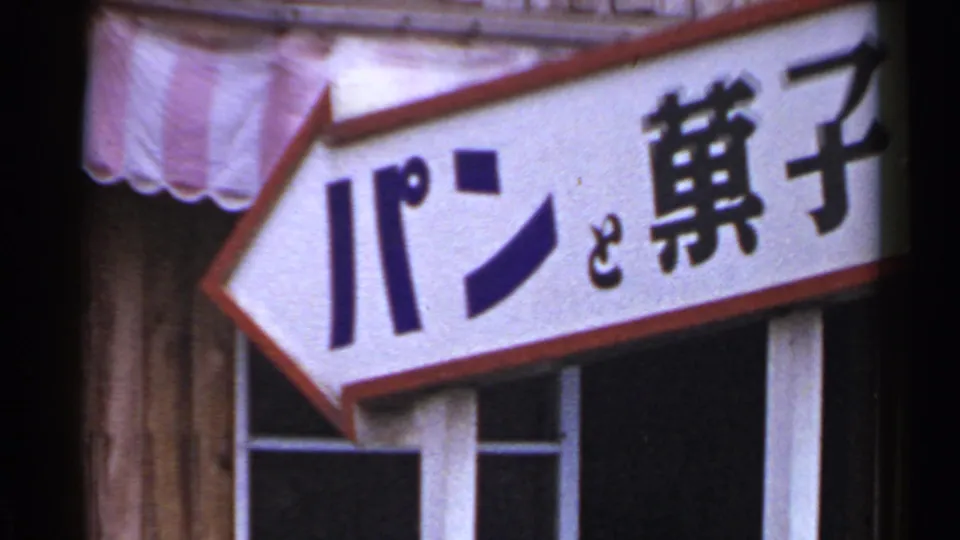 A 1953 Japan street-detail frame with a red-bordered directional sign bearing Japanese characters below the post office wall.