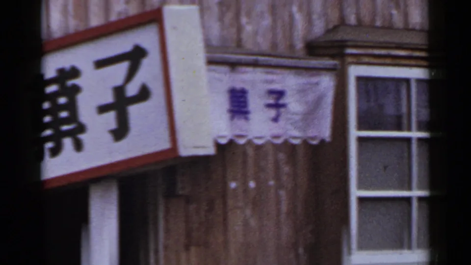 1953 Japan storefront detail with Japanese characters, a small cloth banner, wood siding, and a white-framed window.