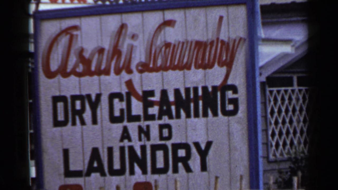 1953 Japan 8mm still showing more of the laundry sign face, including black block lettering and a diamond-patterned window grille.