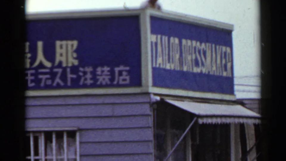 1953 Japan frame showing a corner storefront sign with Japanese text and the English words TAILOR DRESSMAKER above an awning.