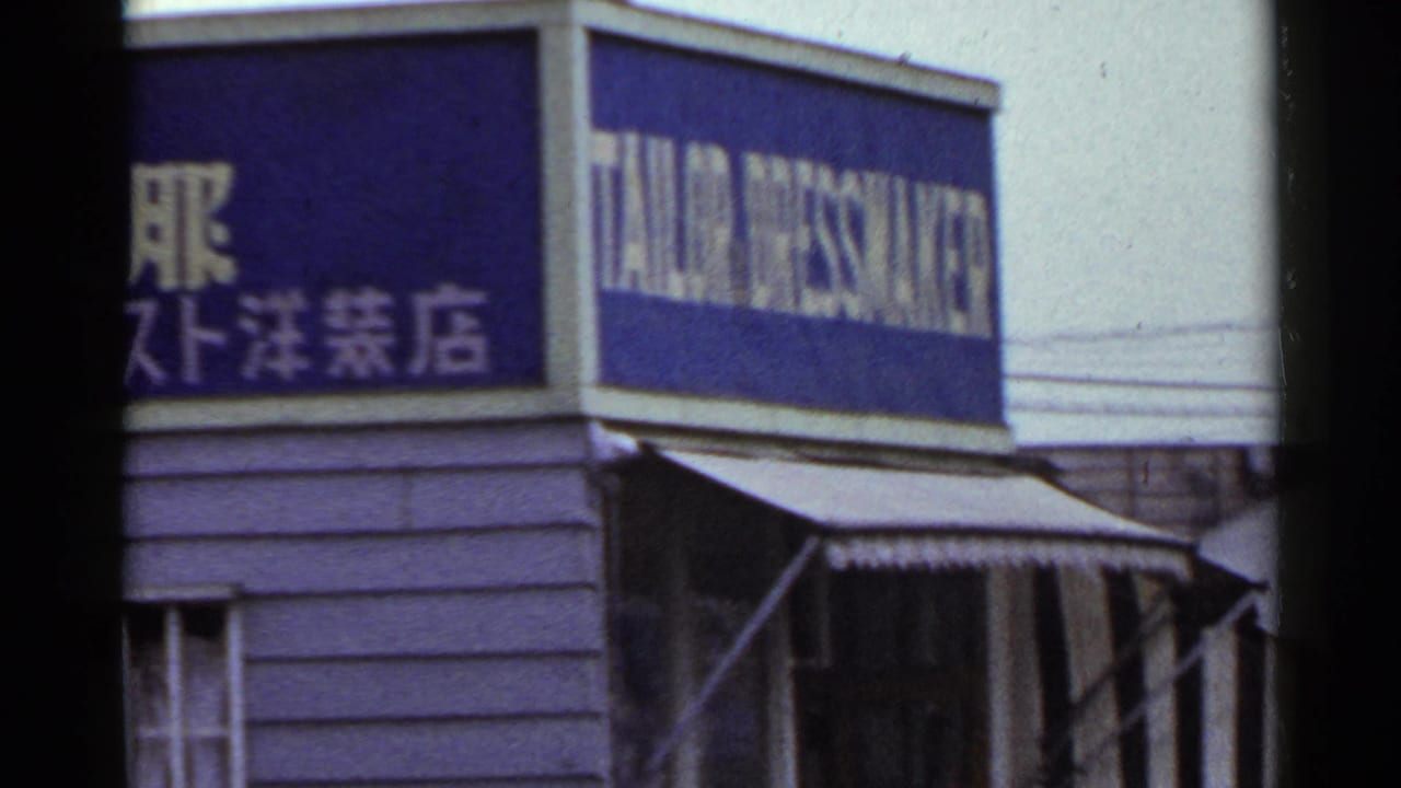 1953 Japan home-movie frame showing a blue tailor and dressmaker storefront sign with Japanese text and a striped awning.