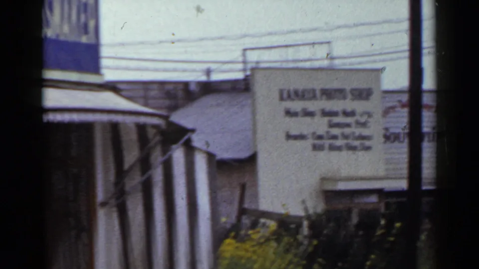 The pan reaches a Kanaya Photo Shop sign near the tailor storefront, with shrubs and shopfronts visible in 1953 Japan footage.