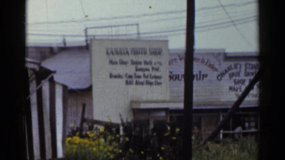 1953 Japan home-movie frame showing Kanaya Photo Shop signage, adjacent small business signs, poles, shrubs, and overhead wires.