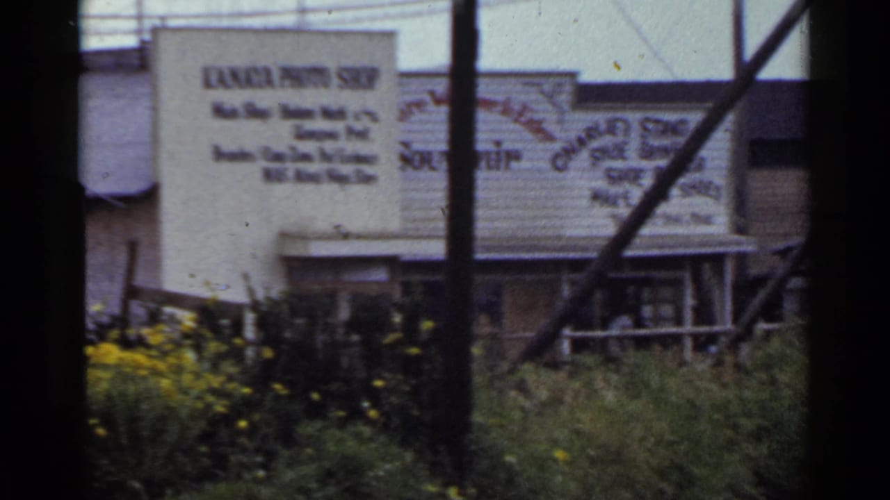 1953 Japan 8mm frame showing weathered commercial buildings, painted signs, poles, bushes, and yellow flowers along a roadside.
