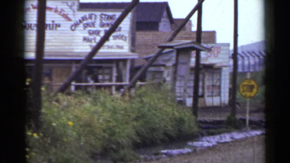 1953 Japan 8mm frame showing a storefront entrance, painted signs, thick roadside bushes, utility poles, and a yellow stop sign.