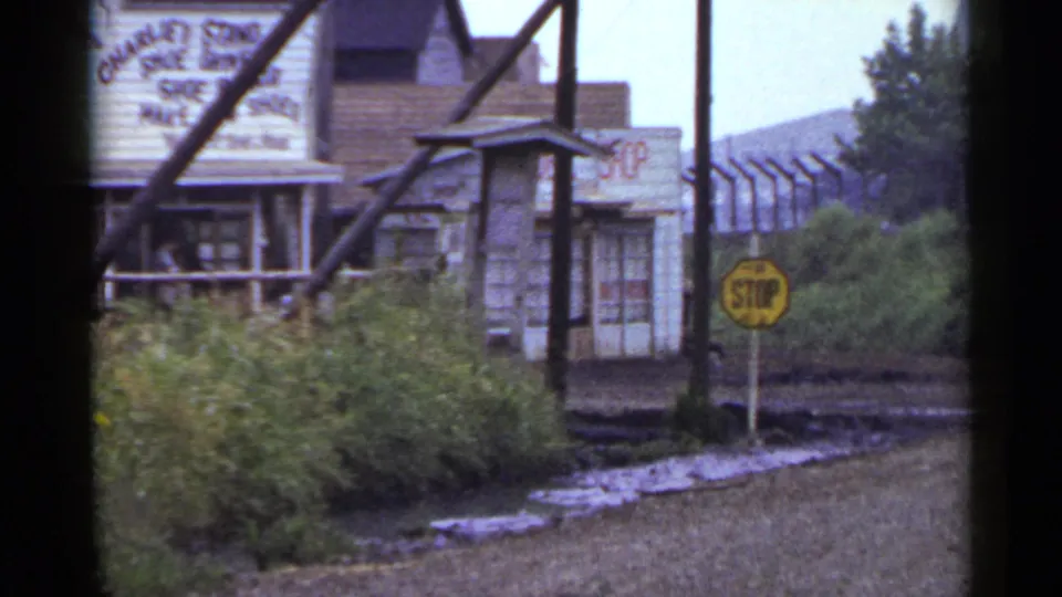 1953 Japan frame with a yellow stop sign near a wet roadside, overgrown greenery, utility poles, and weathered commercial buildings.