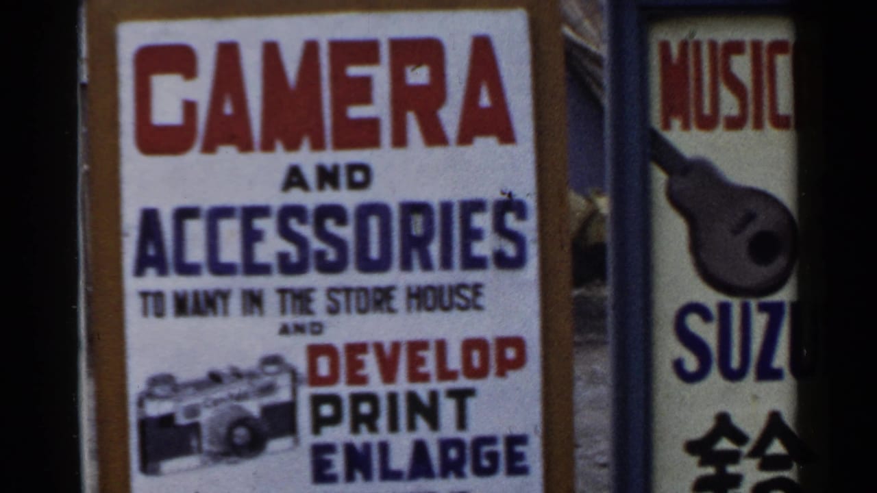 1953 Japan 8mm frame with a camera-service sign and neighboring vertical sign partly visible at the edge of the storefront view.