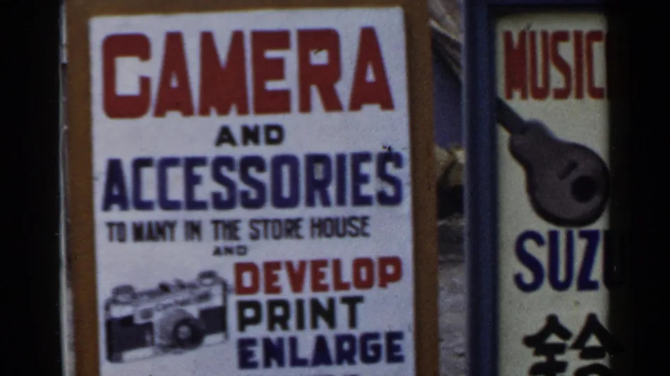 1953 Japan 8mm frame with a camera-service sign and neighboring vertical sign partly visible at the edge of the storefront view.