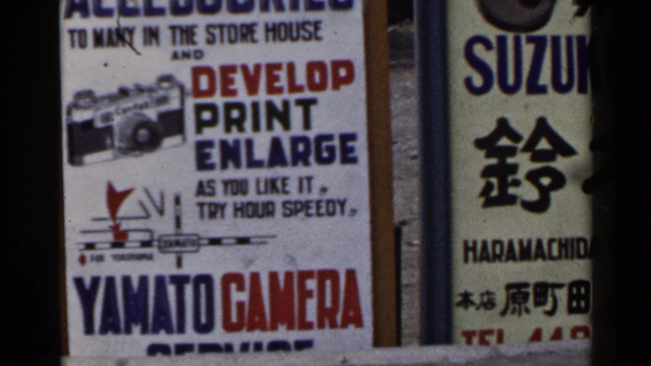 1953 Japan close-up of camera shop advertising, with English service text and Japanese signage partly divided by a dark foreground strip.
