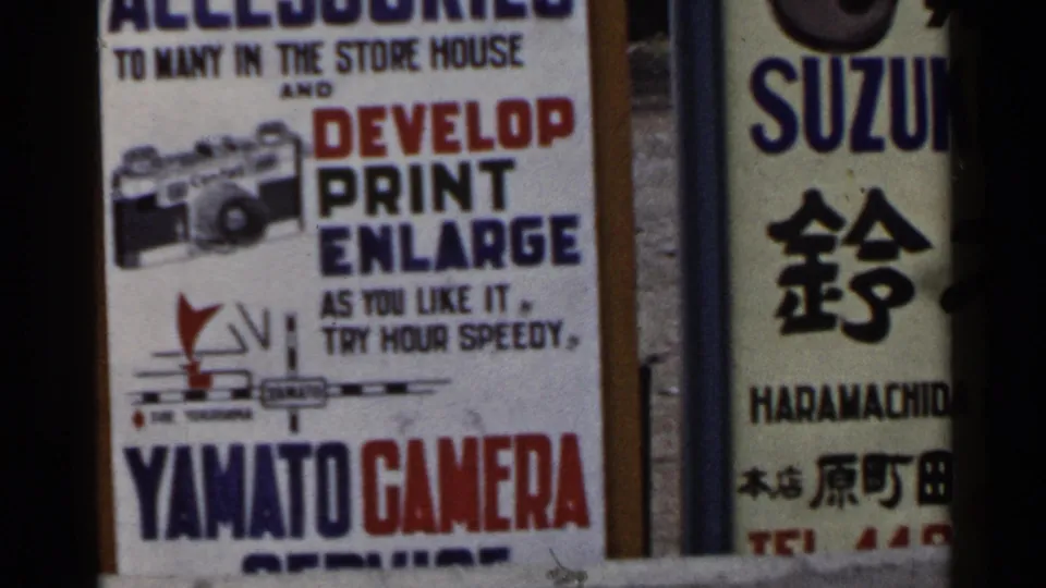 1953 Japan frame dominated by a Yamato Camera advertisement offering photo processing services, with Suzuki and kanji signage alongside.