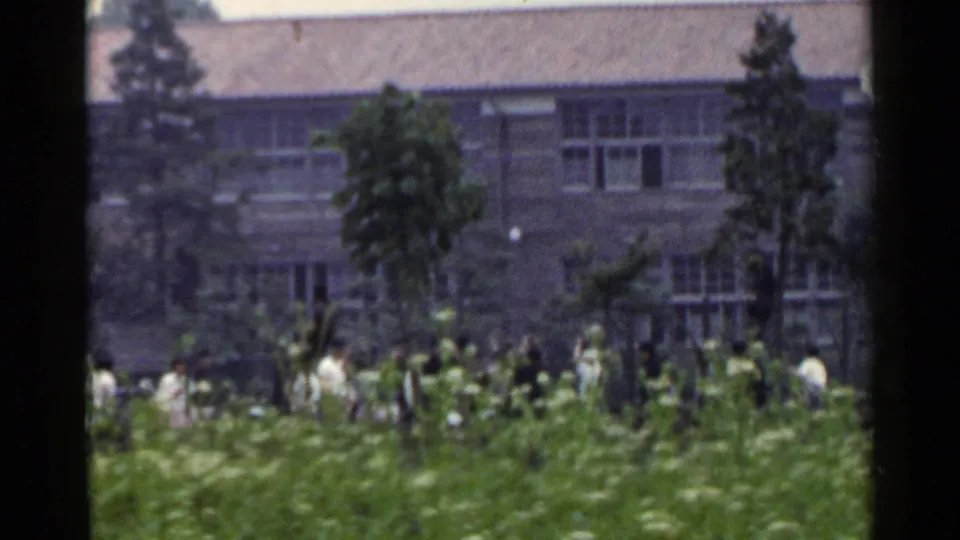 1953 Japan frame showing several distant people near grass and trees before a low building, with the edge of signage or film frame at left.