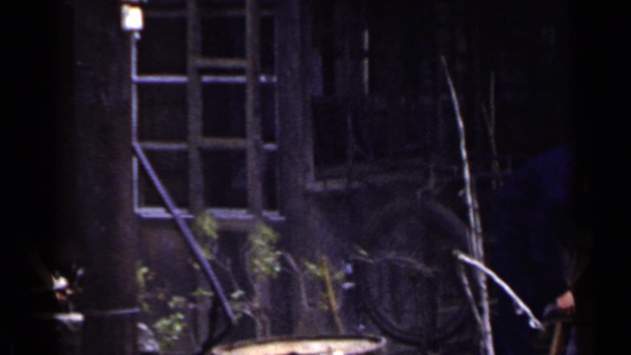 Man partly obscured beside a weathered building, bicycle wheel, branches, and window frames nearby.