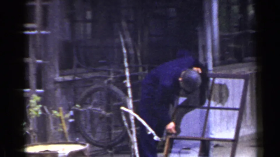 A man in a dark blue jacket and cap bends over a detached window frame outside a weathered building in Japan, 1953.