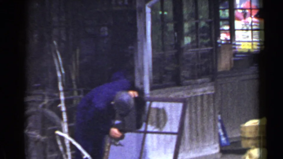 The worker bends over the frame near a shopfront or entry area, with weathered siding and a lit window behind him.