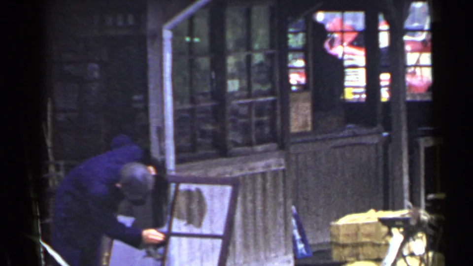 A man remains bent over the leaning window frame beside weathered windows, a broom, and bundled or stacked items in the town setting.