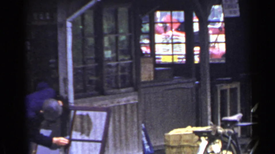 1953 Japan home-movie frame showing a wooden shop or station front, glass windows, posted notices, a bicycle, and a person at the edge.
