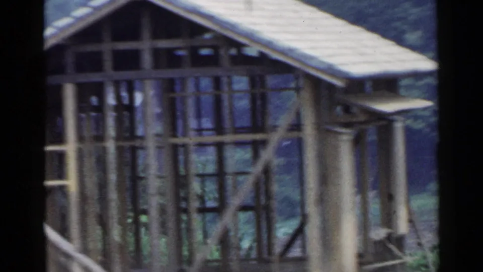 1953 Japan frame showing a wood-framed structure with a tiled roof, open framing, and green hillside beyond.