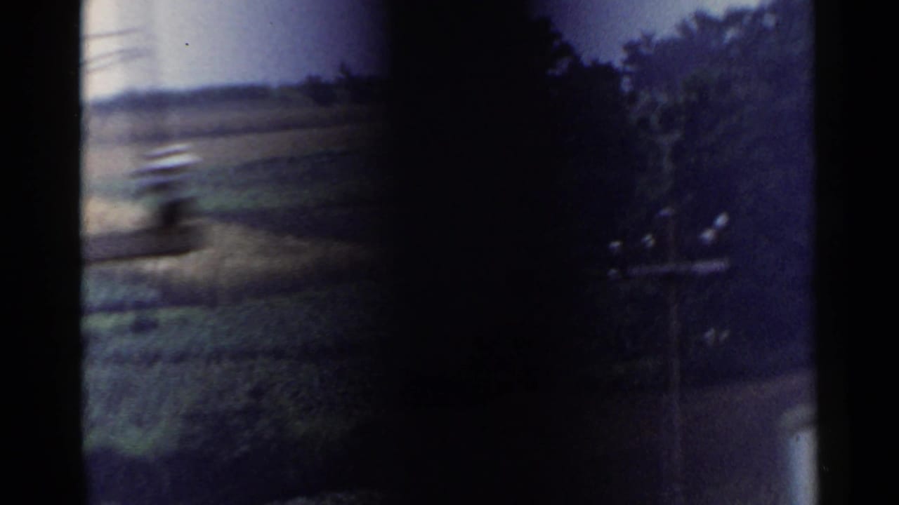 Soft 8mm frame of Japanese countryside with hillside fields, dark gate edges, and trees along the right side.