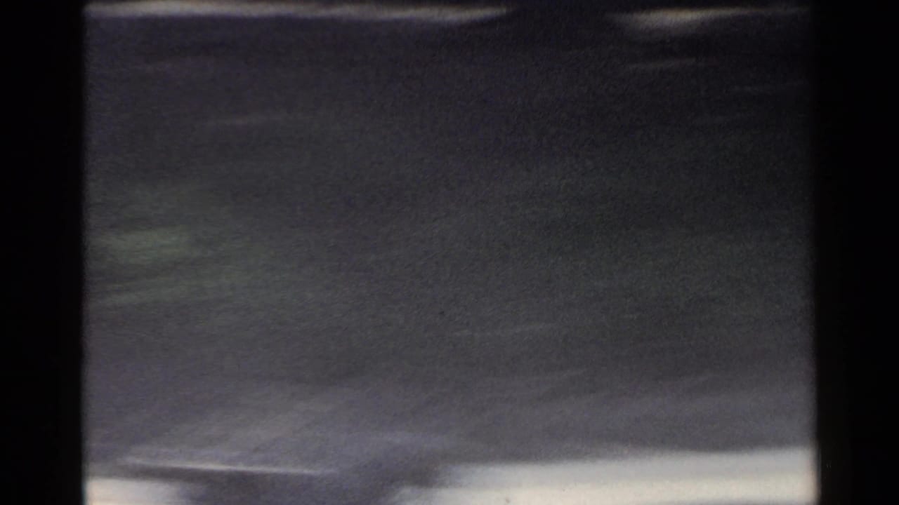 1953 Japan 8mm frame with dark vehicle framing, pale road edge, and blurred roadside scenery passing at speed.