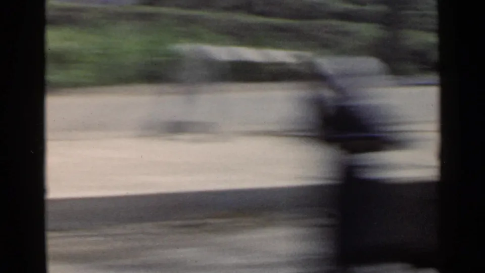 1953 Japan vehicle POV with a low roadside wall, trees, pale pavement, and dark vertical foreground shape blurred by speed.