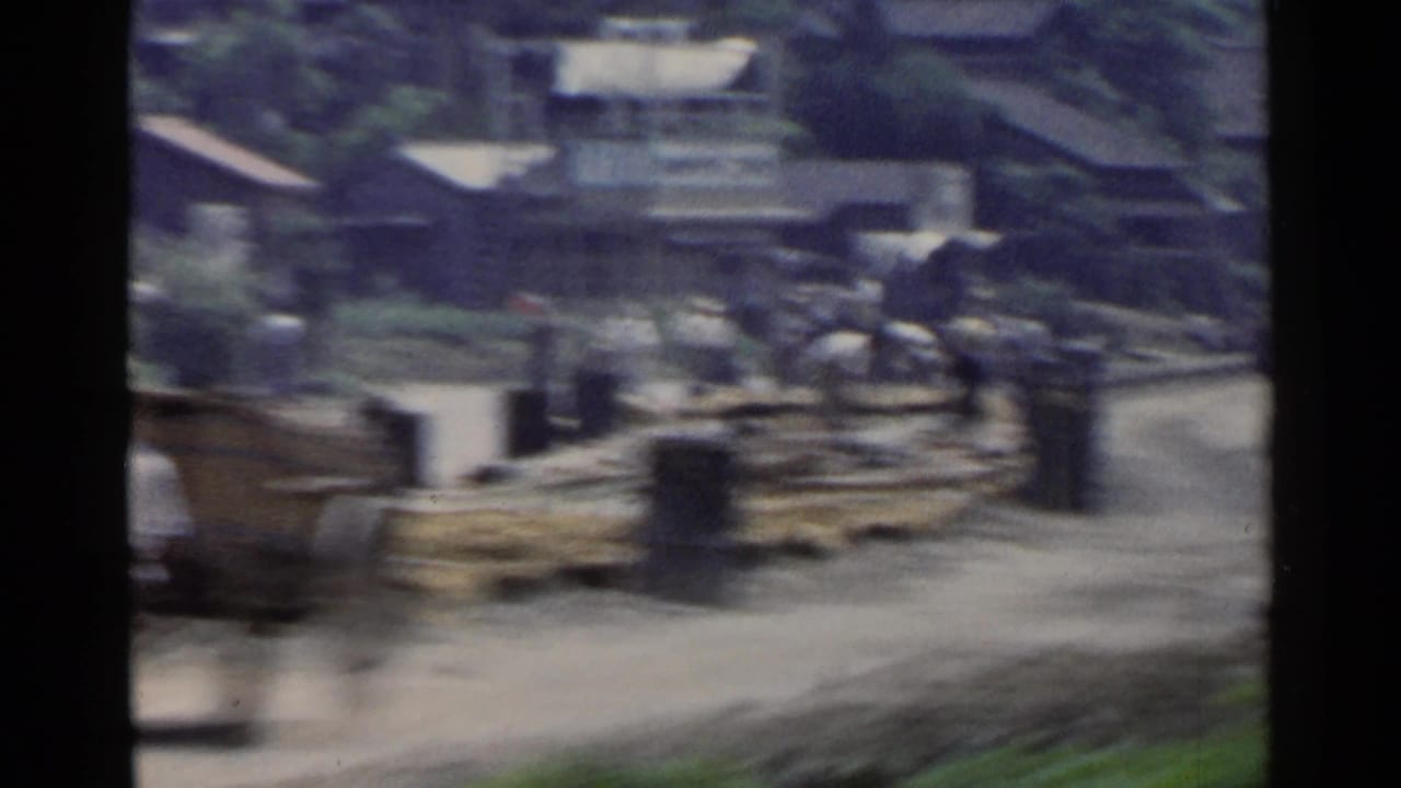 Blurred road and industrial area with buildings, logs, and greenery in the background.