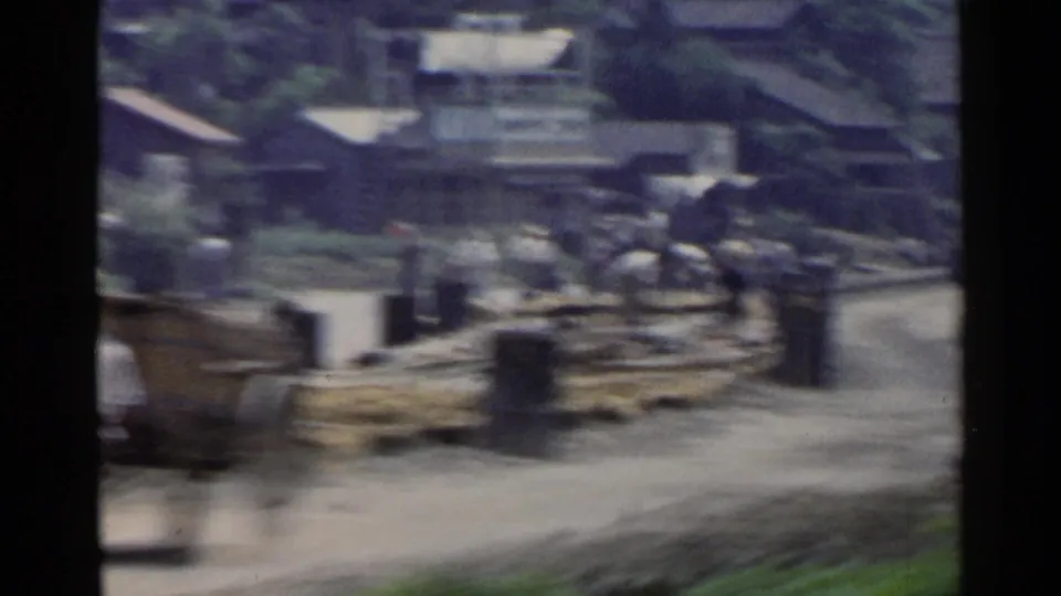 Motion-blurred 1953 Japan street view toward an industrial yard with buildings, stacked timber, and surrounding greenery.