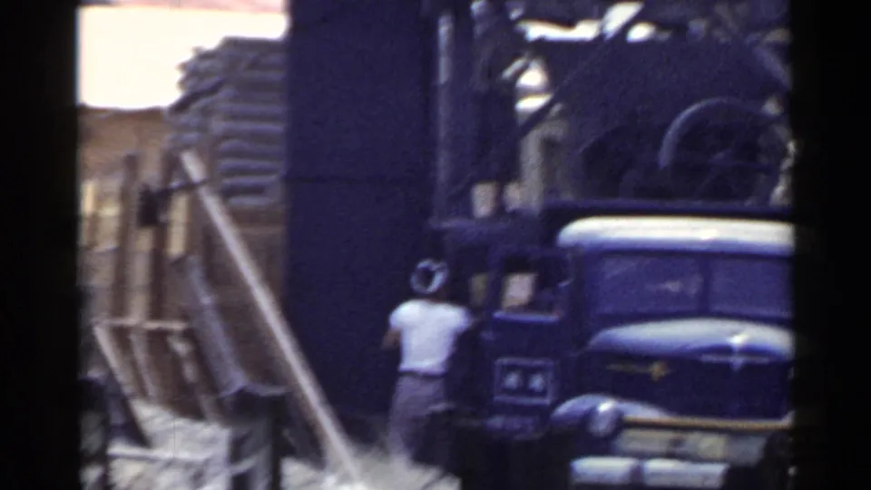 1953 Japan home-movie frame showing a worker near a vintage truck, stacked timber, and a rough wooden loading area.