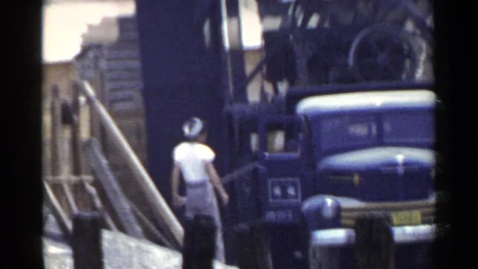 In 1953 Japan, a worker in a light shirt walks past a blue truck beneath a large industrial structure.
