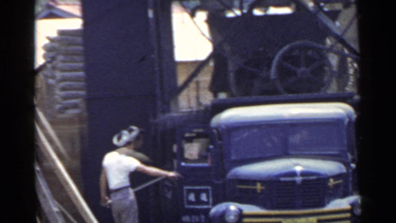 Worker in a white shirt and cap stands beside a blue truck near an industrial structure.