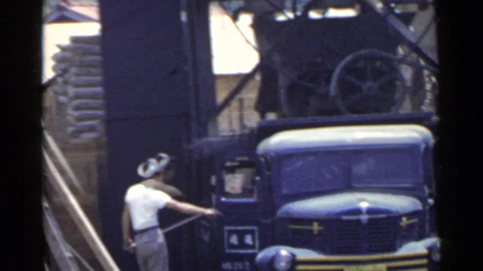 1953 Japan home-movie frame showing a worker beside a blue truck near a dark industrial structure or service area.