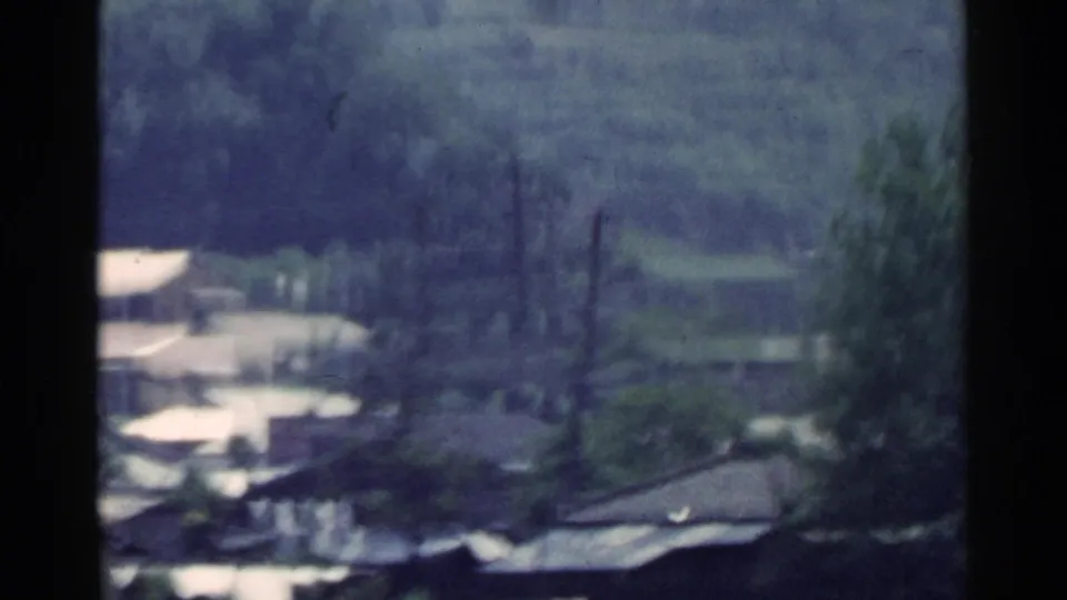 1953 Japan home-movie frame showing a wooded hillside settlement with modest buildings, rooftops, and a pale road or open lane.