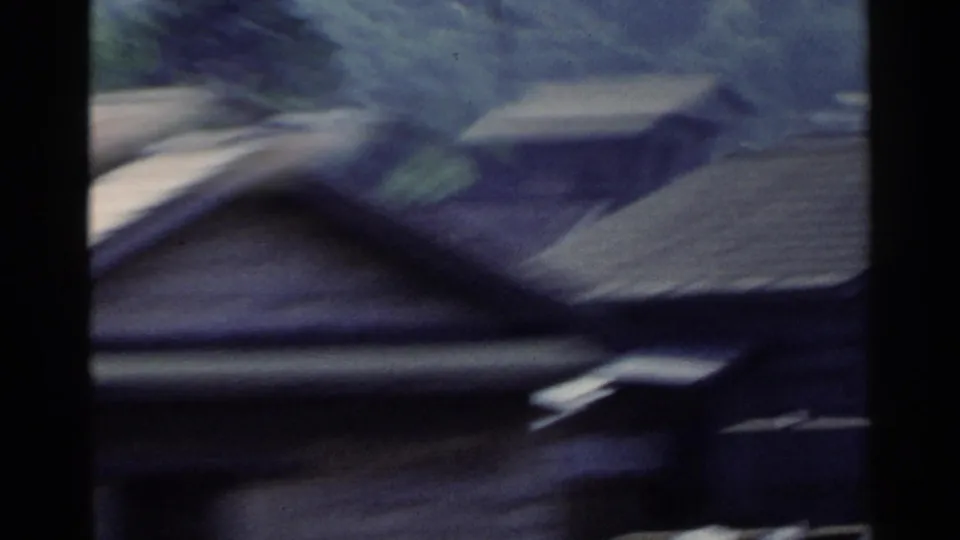 1953 Japan home-movie frame showing a blurred vehicle-window view past rooftops, dark borders, and indistinct urban structures.