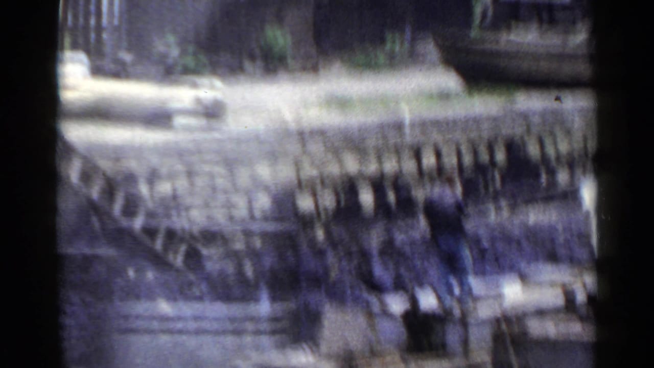 1953 Japan frame with a blurred urban view, showing fencing, pale ground, low structures, and heavy motion smear.