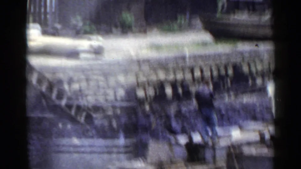 1953 Japan frame with a blurred urban view, showing fencing, pale ground, low structures, and heavy motion smear.