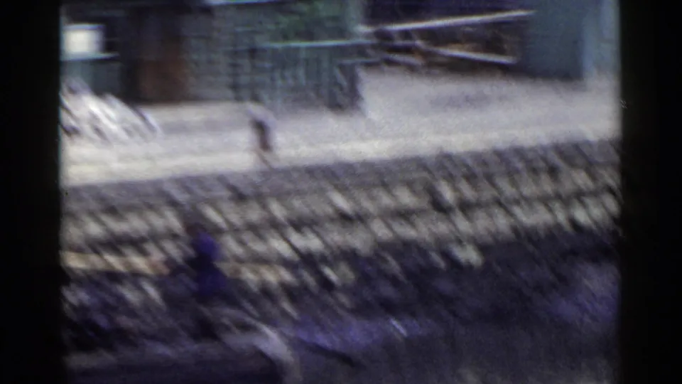 1953 Japan frame showing a shaky moving-vehicle view across fences, open ground, and low buildings with purple-blue film cast.