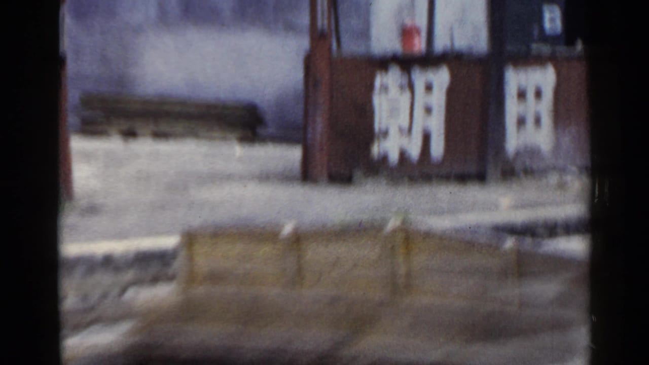 Moving train view in Japan, 1953, with a brown wall carrying bold white Japanese characters and low barriers in the foreground.