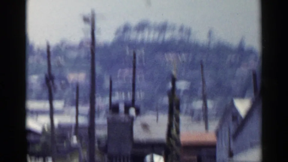 1953 Japan home-movie view of rooftops and utility poles beyond the train route, with a hazy hillside or tree line in the distance.