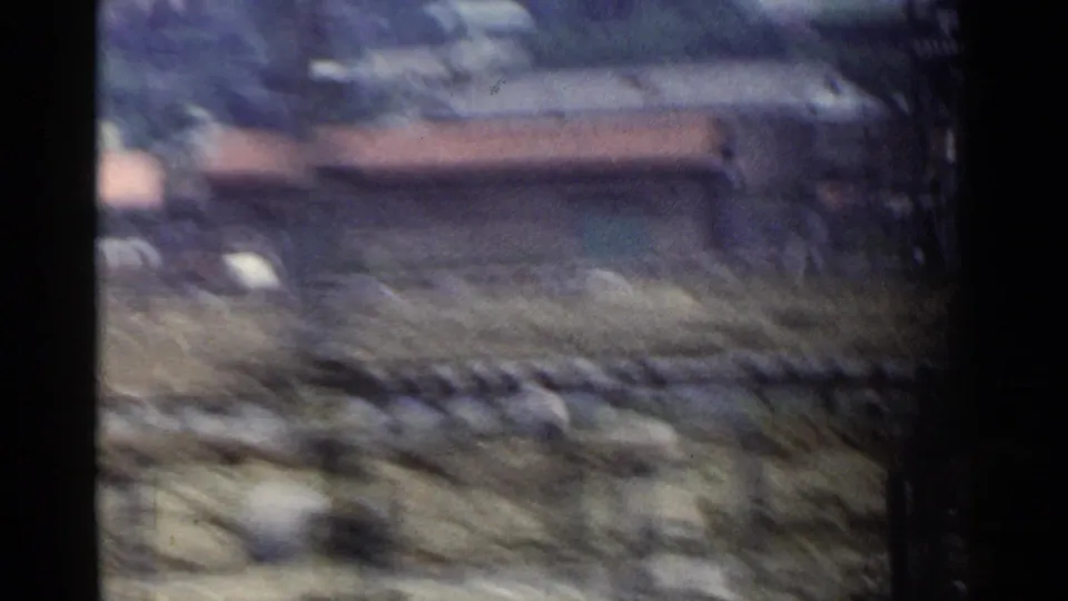 1953 Japan 8mm frame showing blurred ground, a low roofline, haze or smoke, and distant rural structures from motion.