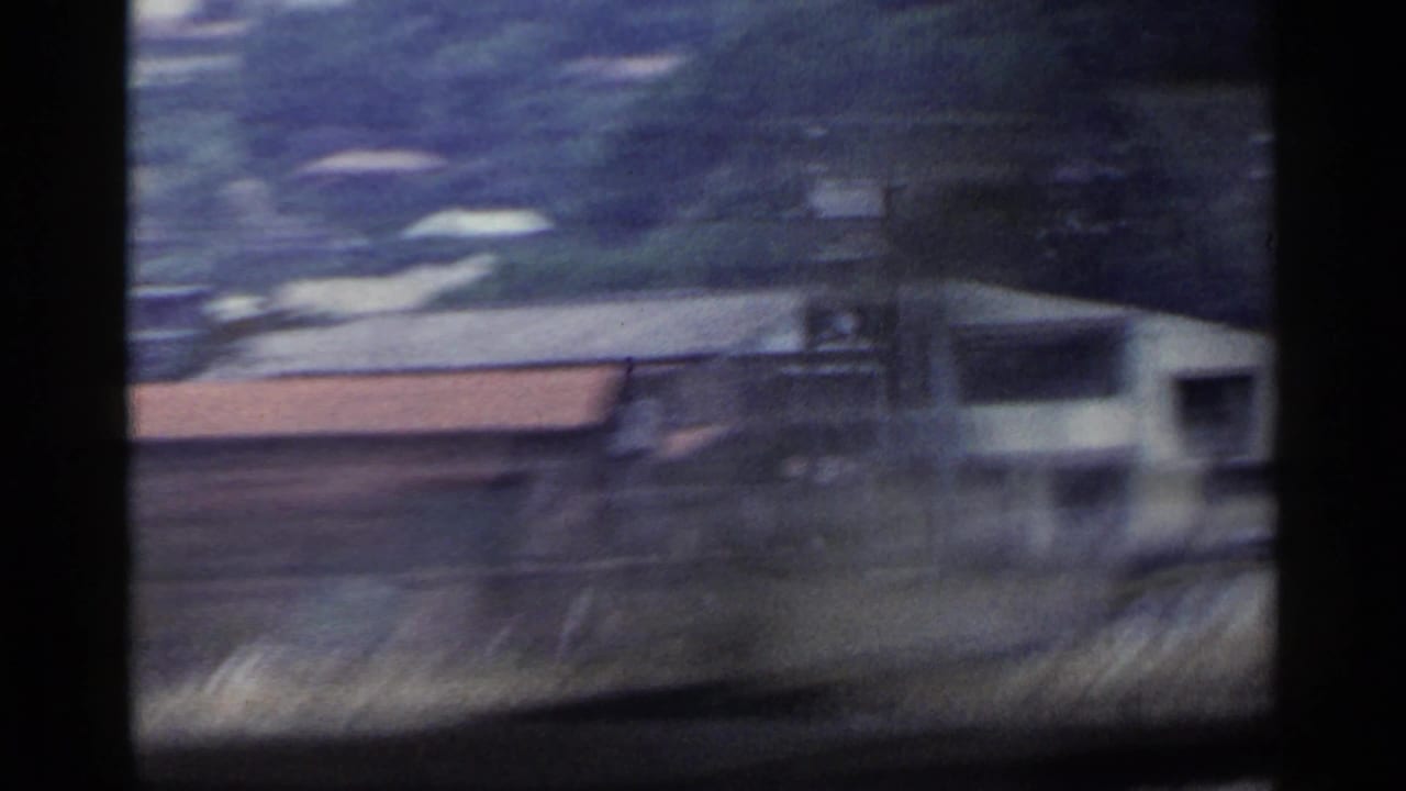 Blurred view from a moving train past rural buildings and trackside ground.