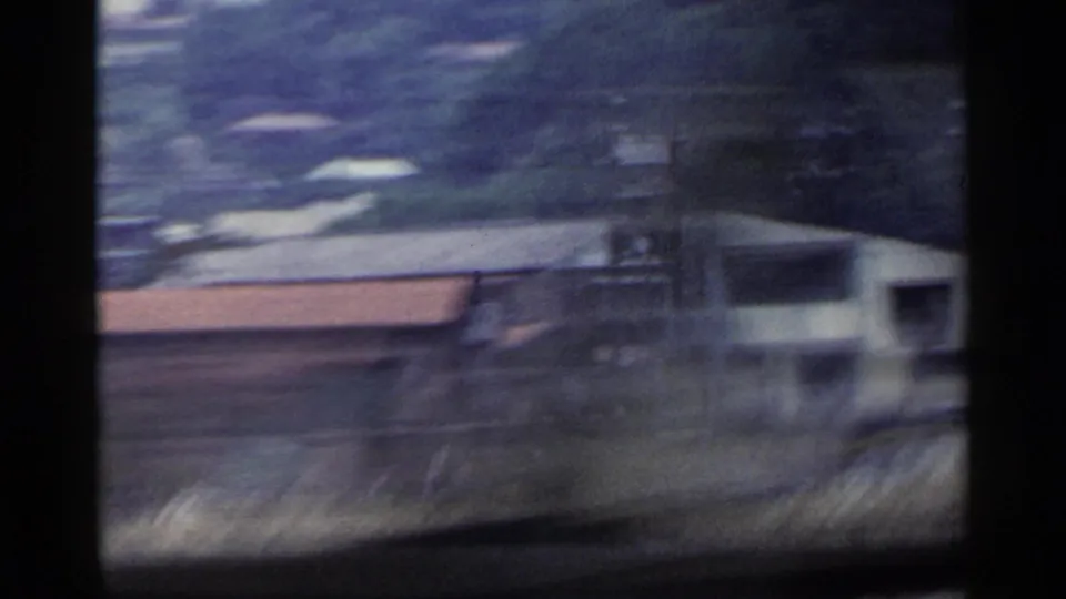 1953 Japan home-movie view from a moving train, with blurred trackside buildings, low roofs, and hillside vegetation beyond.