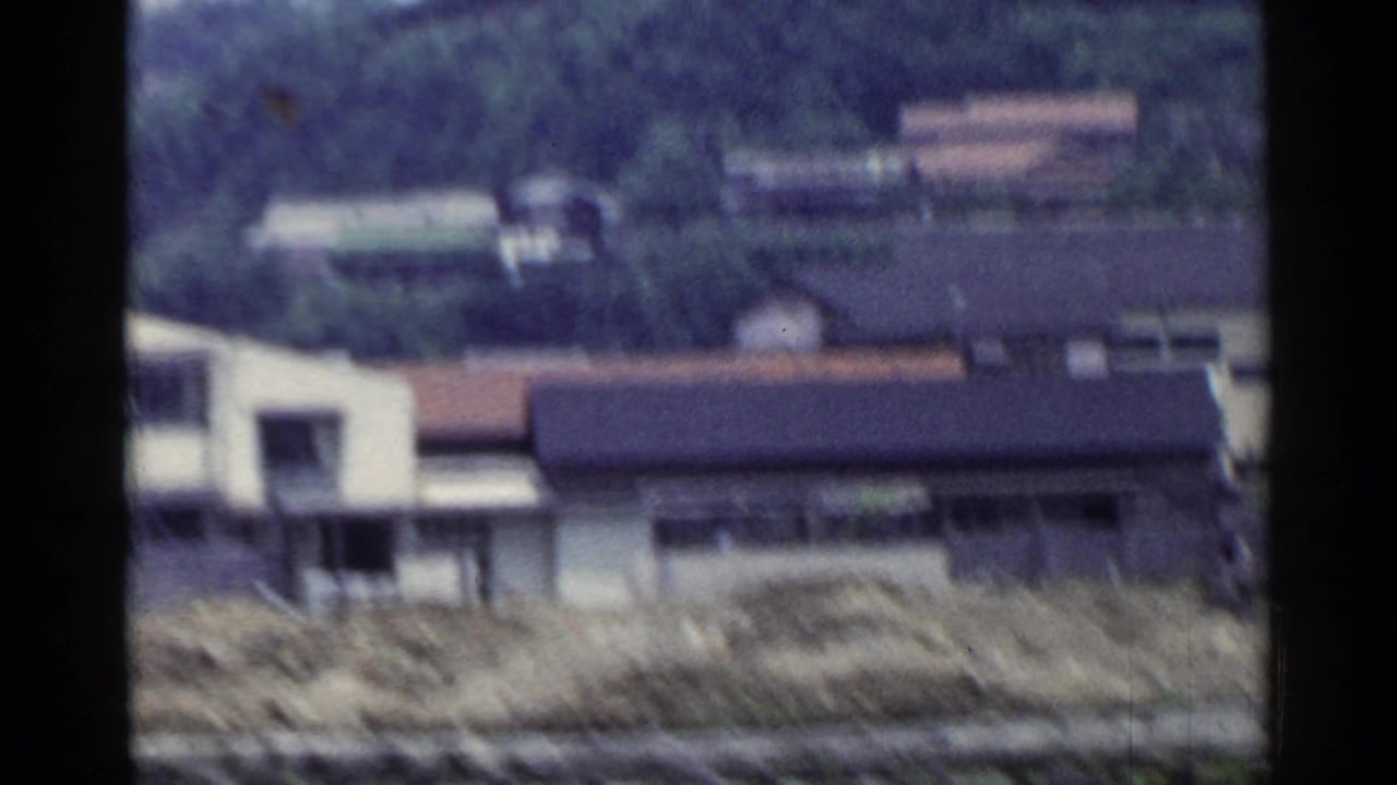 Rural Japanese buildings and a wooded hillside appear through a moving train view in this 1953 8mm frame.