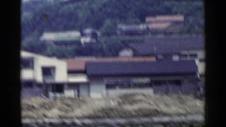 Rural Japanese buildings and a wooded hillside appear through a moving train view in this 1953 8mm frame.