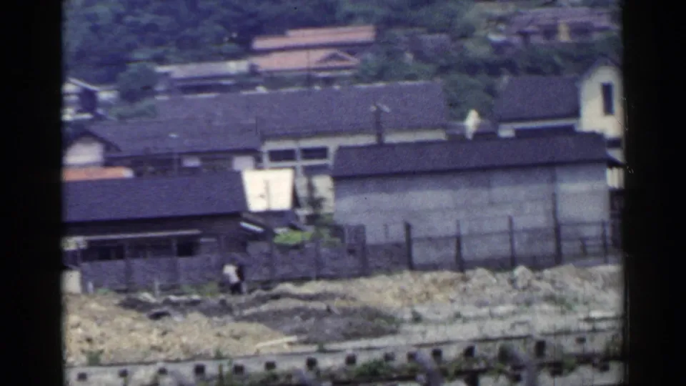 A 1953 Japan rail-side view shows station buildings, rough ground near the tracks, and a distant person walking.