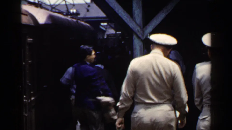 Passengers, including white-uniformed men and a person in a blue jacket, move beside a train at a Japanese station in 1953.