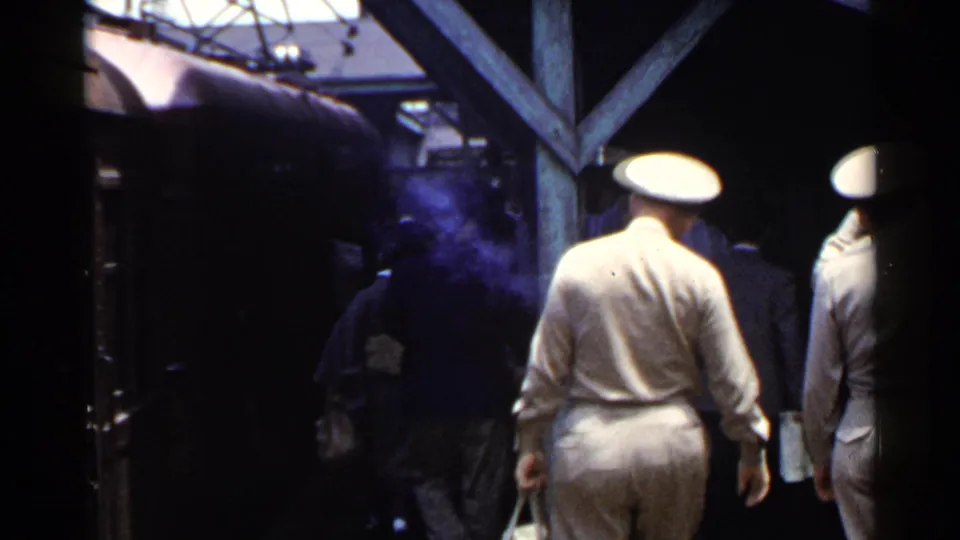 In 1953 Japan, civilians and men in military-style caps move along a crowded train platform under a wooden canopy.