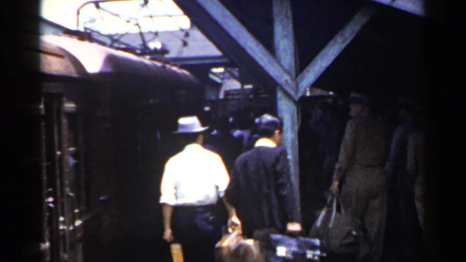 A station platform in 1953 Japan shows civilians in hats and suits walking with luggage near the train and canopy posts.