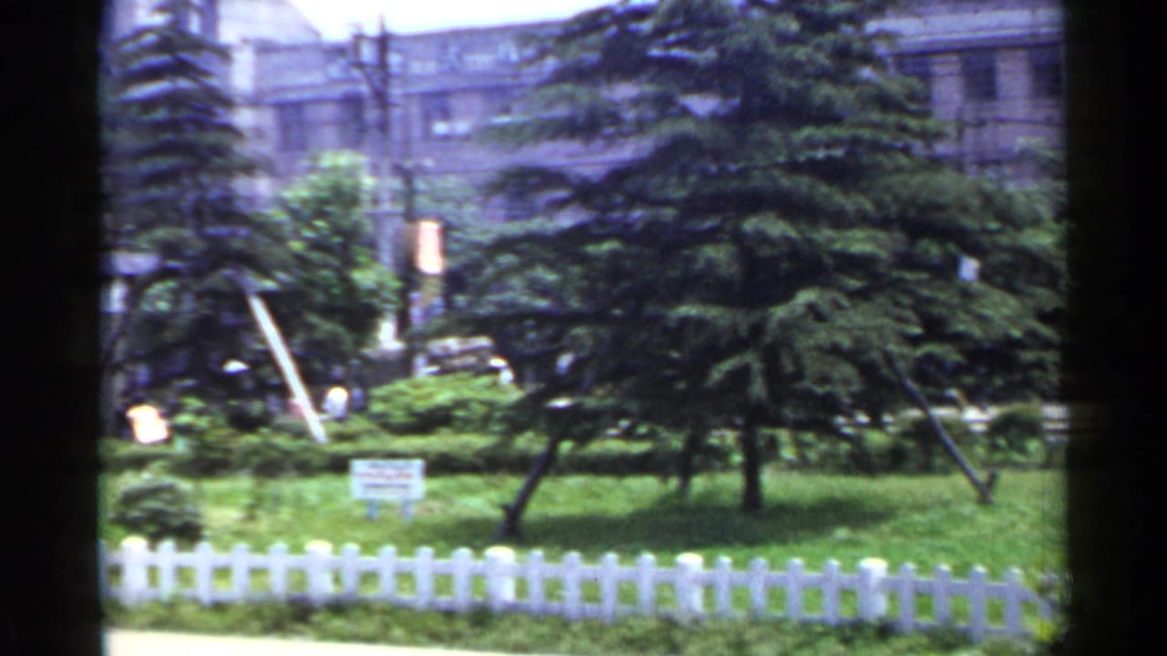 1953 Japan 8mm frame showing a small landscaped park, white picket fence, evergreen trees, and a building with Japanese signage in the background.