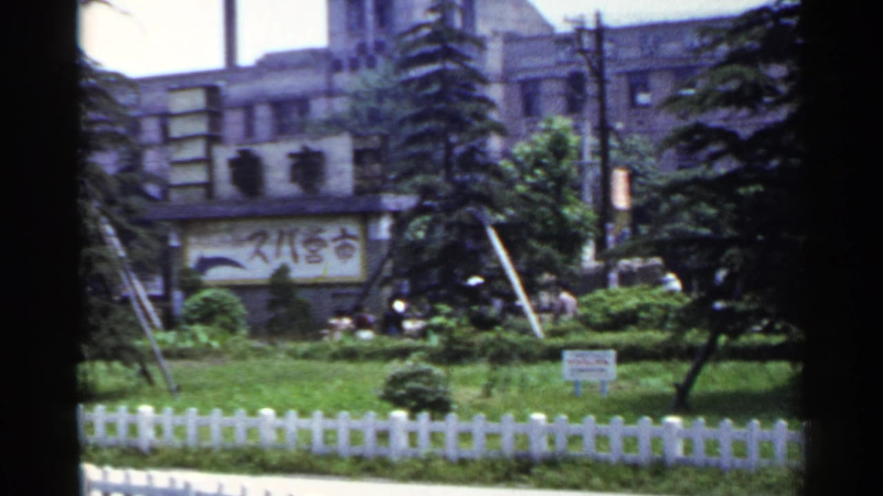 1953 Japan 8mm frame with fenced park landscaping, evergreen trees, overhead wires, and a background building carrying Japanese signage.